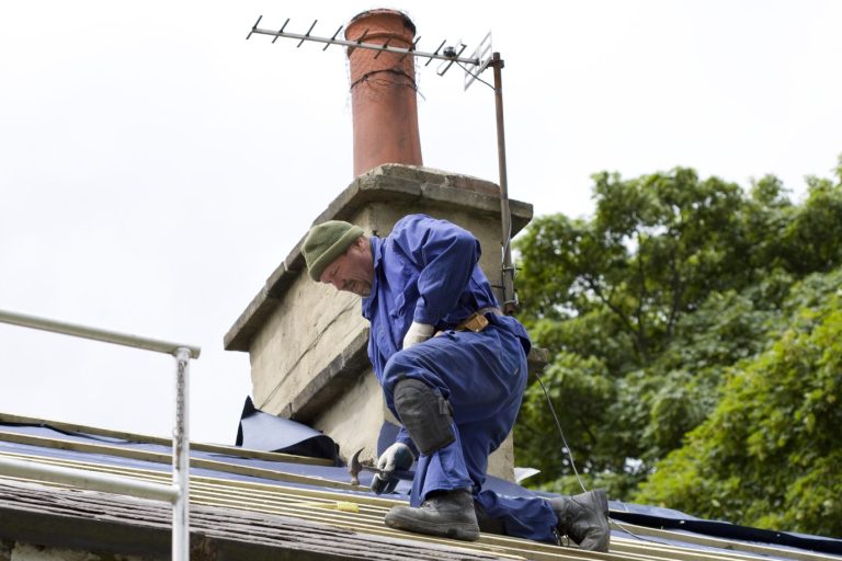 A person in blue overalls climbing on a roof near a chimney and trees completing a roof repair.