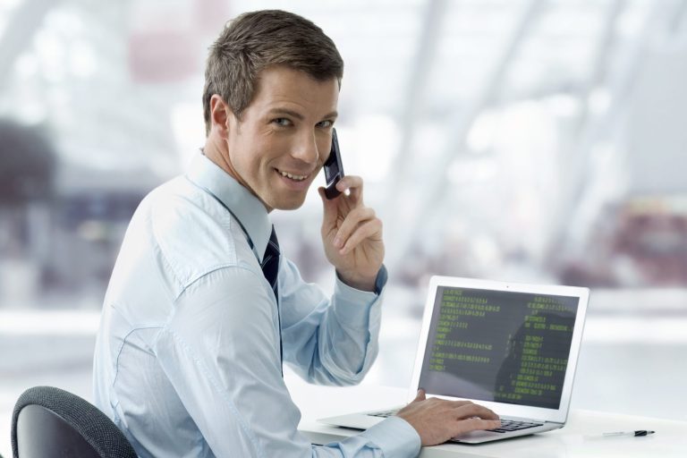 Man in a shirt talks on a phone while working on a laptop.